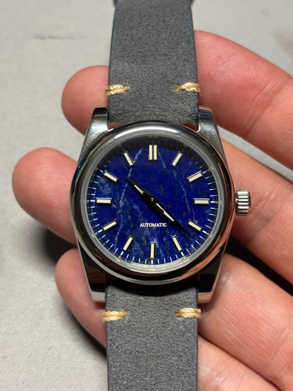 Watch with blue dial and gray strap held in a hand against a neutral background